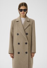 Abrigo de lana beige de doble botonadura con cuello de muesca, botones negros y bolsillos laterales. Lleva puesto un suéter ligero y está combinado con gafas de sol negras.