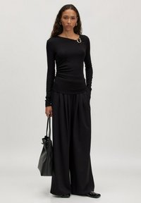 Schwarzes Langarmoberteil mit geraffter Textur und einem dekorativen silbernen Akzent. Kombiniert mit weiten schwarzen Hosen und einer schwarzen Tragetasche.