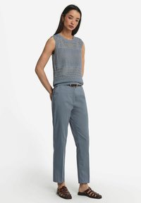 Ljusblå mönstrad stickad ärmlös topp i kombination med ljusblå byxor och bruna sandaletter med remmar. Neutral bakgrund.