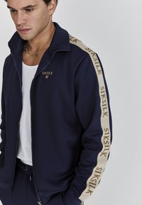 Námornícky zipsový kabátik vyrobený z hladkej tkaniny, s kultovými zlatými akcentmi a svetlobežovou logovou prúžkou pozdĺž rukávov. Slim fit dizajn.