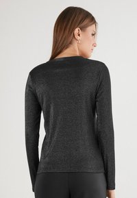 Žena s dlouhými hnědými vlasy, která má na sobě stříbrné kruhové náušnice a přiléhavý černý třpytivý top s dlouhým rukávem, viděná zezadu.