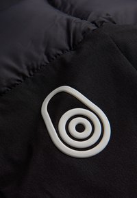 Svart tyg med en texturerad logotyp i vitt, formad som en abstrakt oval med koncentriska cirklar, detaljerad sömnad runt kanterna.