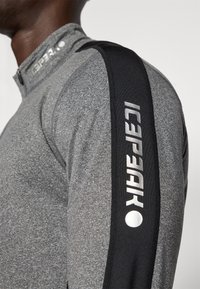 Šedý sportovní zip-up top s černým akcentem na rameni. Obsahuje reflexní branding. Texturovaná tkanina s přiléhavým střihem a vysokým límcem.