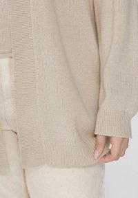 Beige gestrickte Strickjacke mit einem gerippten Kragen und Bündchen, in lockerer Passform und mit weicher Textur, dargestellt mit einer Hand, die den Saum hält.