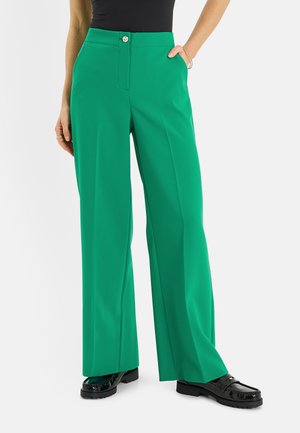 Mujer con pantalones anchos de color verde brillante, top negro y mocasines negros brillantes, con una mano en el bolsillo y una pulsera dorada.