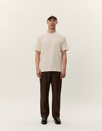 Beige rövidujjú ing, barna nadrág, fekete cipő, és egy világos színű sapka logóval. A modell egy egyszínű háttér előtt pózol.