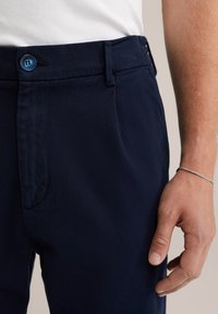 Bordo krāsas bikses ar gludu tekstūru, ar zilu pogu un sānu kabatām. Roka novietota uz jostas detaļām.