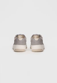 Šedé semišové tenisky s nízkym profilom, béžovou gumovou podrážkou a modrým vnútorným podšívaním. Na zadnej strane sa nachádza logo Reebok na pätnom pútku.