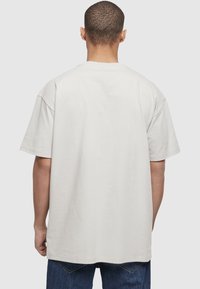 Mand iført ensfarvet lysegrå oversized t-shirt og blå jeans, set bagfra mod en lysegrå baggrund.