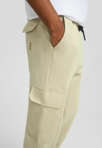 Ljust beige cargo-byxor med en slät textur, sidofickor och midja med dragsko, med en liten logga på vänstra fickan.