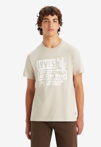 Camiseta de algodón en color beige claro con un atrevido texto gráfico en blanco y diseño de cactus. Mangas cortas y escote redondo.