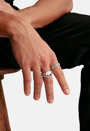 Linke Hand ruht auf dem Knie, trägt zwei silberne Ringe: einen mit Totenkopfdesign am Mittelfinger und einen gemusterten Ring am Zeigefinger.