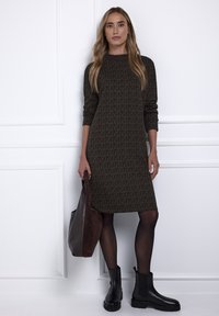 Sukienka dzianinowa w ciemnozielonym kolorze z geometrycznym wzorem, z długimi rękawami i lekko dopasowanym krojem. Połączona z czarnymi botkami na kostkę i brązową torbą typu tote.