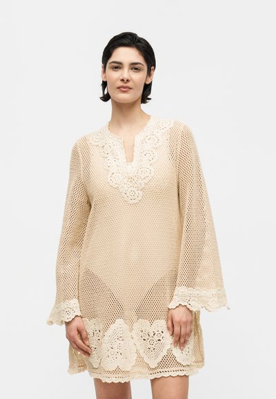 Femme portant une robe beige en crochet à manches longues avec un bord en dentelle florale au niveau de l'encolure, des poignets et de l'ourlet, debout devant un fond blanc.