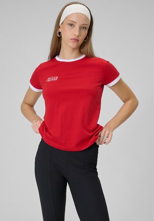 Kobieta w czerwonym T-shircie z białym obszyciem i czarnych spodniach, pozująca z rękami na biodrach, nosząca białą opaskę na głowę i srebrne kolczyki koła.