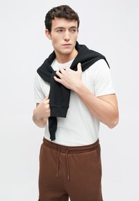 Hvid t-shirt, brune joggingbukser og sort hættetrøje draperet over skuldrene. Glat stof og løs pasform. Model poserer med den ene hånd på hættetrøjen.