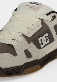 Κοντινή λήψη ενός μπεζ και καφέ παπουτσιού DC skate με λευκά κορδόνια και το λογότυπο DC στη πλευρά και στη γλώσσα.