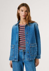 Leren denim jack in lichtblauw met knoopsluiting, twee voorkeurszakken en een geborduurd logo-detail. Draagt over een gestreepte rode en zwarte top.