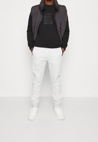 Chaleco negro con cremallera sobre una camiseta gráfica negra, combinado con pantalones de chándal gris claro y zapatillas blancas. Tejido suave, corte casual.