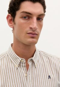 Camicia a righe beige e bianca con colletto button-down, caratterizzata da un piccolo logo ricamato di teschio e tibie sul lato sinistro del petto.
