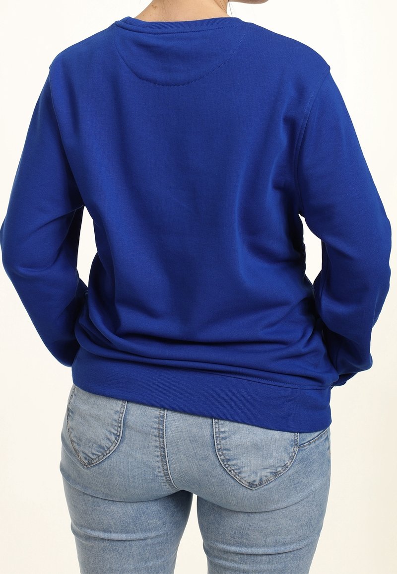 Personne portant une chemise bleue unie à manches longues et un jean en denim bleu clair, vue de dos sur un fond blanc.