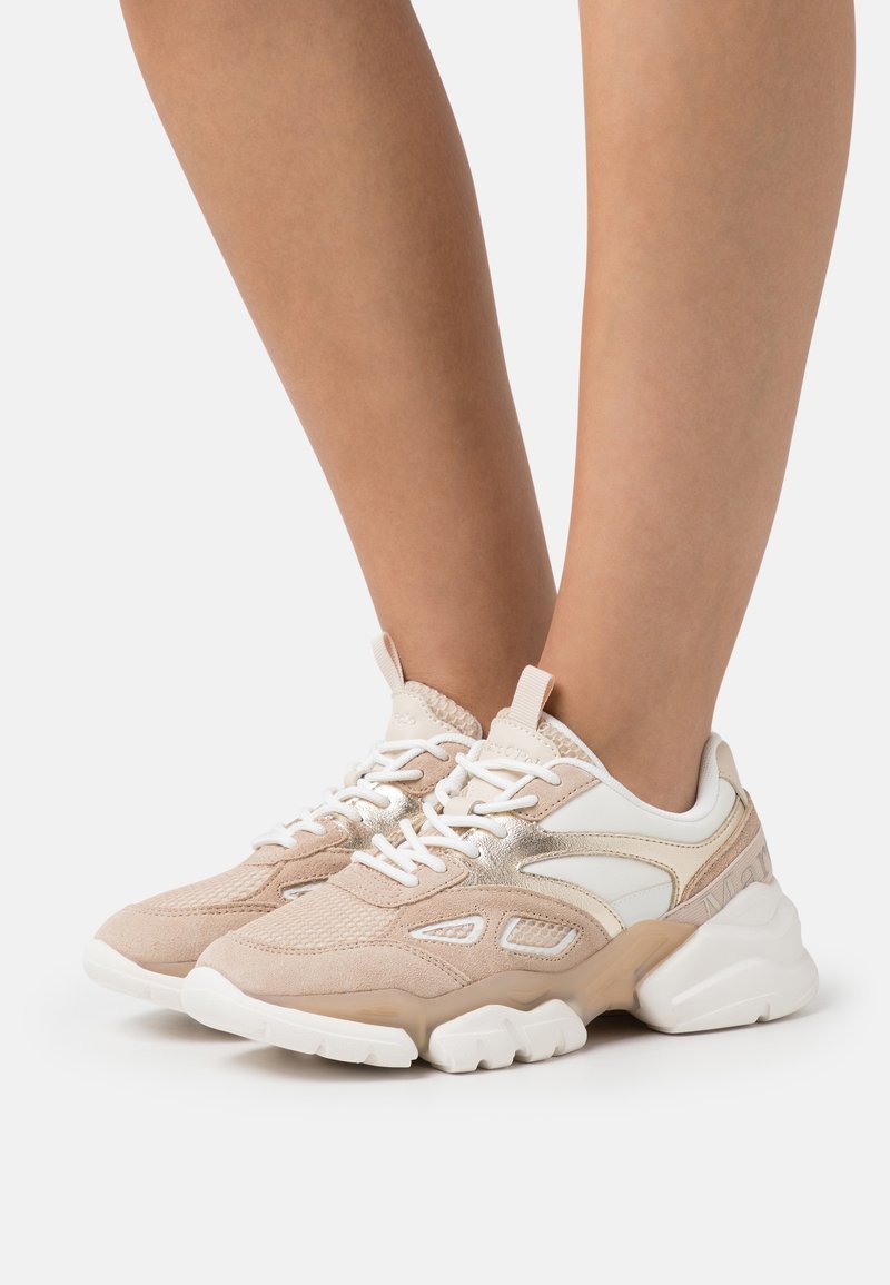 Pieds chaussés de baskets épaisses beige et blanches avec des détails en mesh, en daim et métalliques sur un fond blanc.