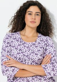 Fioletowa sukienka w kwiaty z białymi i czarnymi kropkami, o rękawach do połowy łokcia i zaokrąglonym dekolcie. Miękka faktura tkaniny.