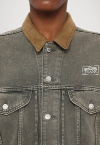 Denim jacket with a brown corduroy collar, silver buttons, and a white logo patch on the chest. The fabric has a faded gray hue and textured surface.