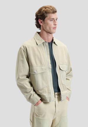Jeune homme aux cheveux bouclés châtain clair portant une veste beige zippée avec de grandes poches par-dessus une chemise foncée, les mains dans les poches du pantalon.