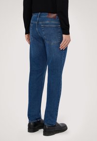 Jeans de mezclilla azul con un corte relajado, bolsillos traseros y un parche de cuero marrón con el logo. El material tiene un aspecto suave y ligeramente texturizado.