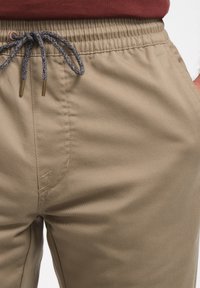 Pantalons en mélange de coton beige avec une taille élastique et un cordon de serrage gris. Le tissu présente une texture lisse et des coutures subtiles.