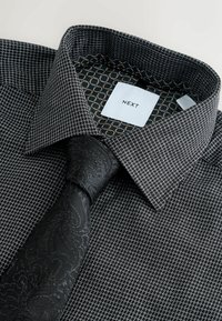 Melnā rakstaina krekls ar strukturētu audumu, ar kontrastējošu heksagonālu rakstu ap kakla daļu un paisley motīvu kaklasaiti. Uz birkas redzama zīmola nosaukums "NEXT."