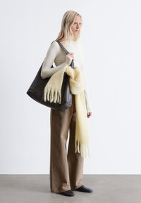 Black leather tote bag with a yellow wool scarf draped over it. Model wears a cream top and brown wide-leg pants, standing on a smooth floor.