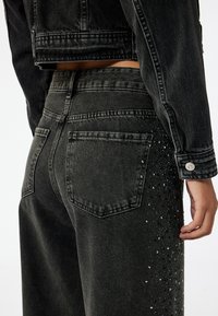 Sorte denimjeans med en afslappet pasform, prydet med nitter på siden. Stoffet har en blød tekstur og et falmet udseende.