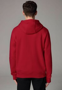 Felpa rossa con cappuccio grande e regolabile, polsini e orlo a coste; tessuto liscio; mostrata di spalle con una vestibilità dritta e senza loghi.