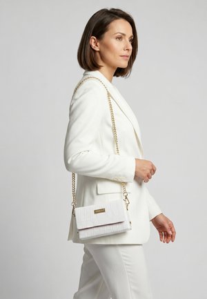 Femme en costume blanc portant un sac à main blanc texturé avec une bandoulière chaîne dorée sur l'épaule, tournée vers la droite sur un fond uni.