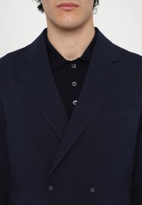 Sacou dubluț cu dungi navy, cu rever în vârf, un buzunar pe piept și închidere cu două butoane; asortat cu un tricou polo negru.