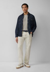 Een blauwe denim overhemd over een grijs gestreept T-shirt, beige broek en witte sneakers met bruine zolen. Heeft zakdetails en knoopsluitingen.