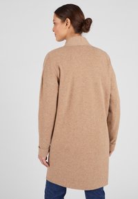 Cardigan lungo in misto lana beige con una vestibilità ampia, colletto arrotolato, spalle scese e tasche applicate; texture morbida e design di lunghezza media.