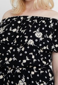 Czarna sukienka opuszczona na ramionach z białym florystycznym wzorem, warstwowymi falbanami i teksturowanym materiałem. Zawiera małe pompony wzdłuż krawędzi.