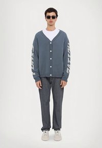 Blauer Cardigan mit geripptem Halsausschnitt, Knopfleiste und weißem Textdesign auf den Ärmeln, kombiniert mit dunklen Jeans und weißen Turnschuhen.