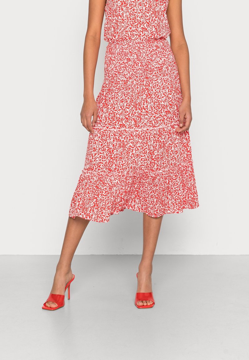 Femme portant une jupe midi fleurie rouge et blanche avec un ourlet à volants et des chaussures rouges à talons hauts ouvertes à l’avant, debout les jambes croisées.