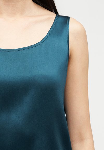 Γυναίκα που φοράει αμάνικο τοπ από σατέν σε τιρκουάζ χρώμα με στρογγυλεμένο ντεκολτέ, σε ουδέτερο φόντο, εμφανίζοντας τον ώμο και το πάνω μέρος του στήθους.
