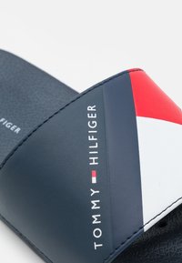 Granatowe klapki z syntetycznej skóry z czerwono-białym ukośnym paskiem. Nadrukowany tekst "TOMMY HILFIGER" na biało. Teksturowana podeszwa.