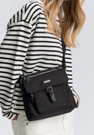 Femme portant un haut à rayures noir et blanc, portant un sac bandoulière noir avec des détails de fermeture éclair et de boucle, pantalon blanc visible.
