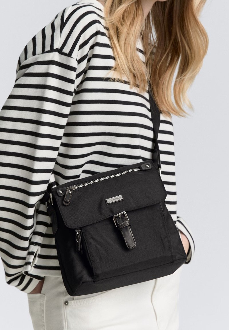 Donna che indossa una maglietta a righe bianche e nere, con una borsa a tracolla nera dotata di zip e dettagli in metallo, e pantaloni bianchi visibili.