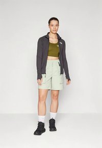 Μαύρο μπουφάν με φερμουάρ, τοπ σε ελιά πράσινο χρώμα, ανοιχτό πράσινο σορτς με τσέπες, μαύρα ορειβατικά μποτάκια, λευκές κάλτσες, στέκεται σε απλό φόντο.