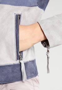 Main dans la poche latérale avec tirette de fermeture éclair argentée visible et manche de veste en daim gris et bleu avec tirette assortie.