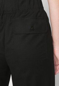 Gros plan sur un pantalon noir avec une poche arrière boutonnée et une taille élastique, montrant une partie du bras d'une personne contre un fond gris.