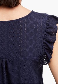 Haut bleu marine avec texture en dentelle à motifs en losange, manches volantées et encolure ronde. Tissu doux avec des détails délicats.